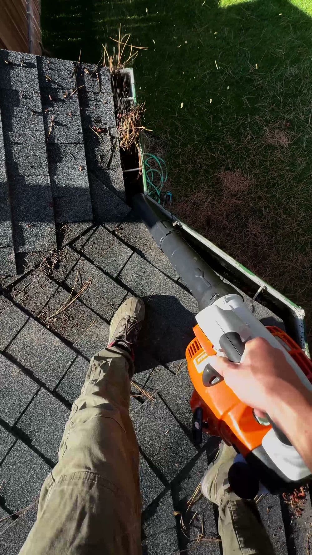 Person using a leaf blower to clear debris from a roof covered in shingles.