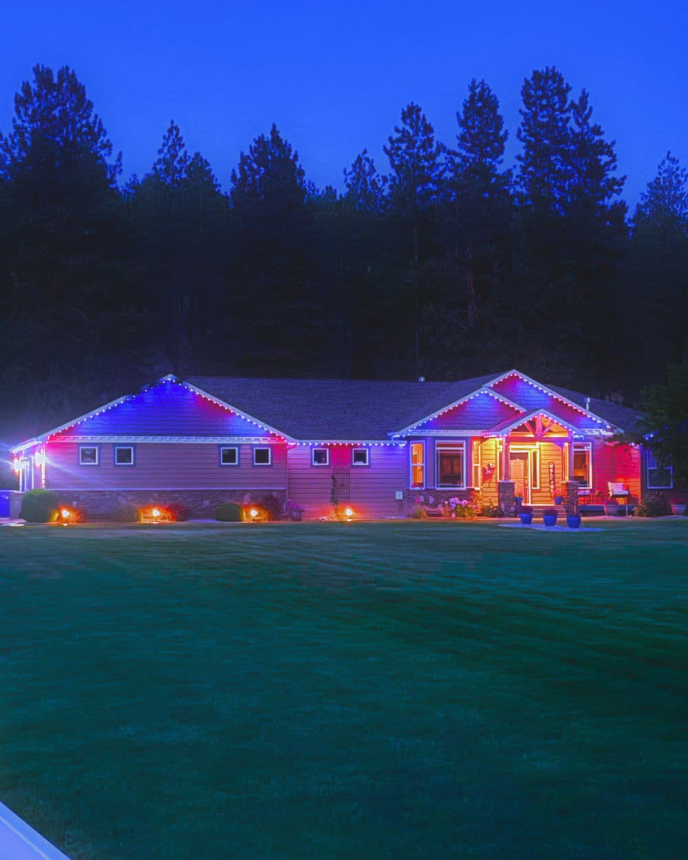 Festively lit home at dusk, surrounded by tall trees and green lawn, showcasing holiday spirit.