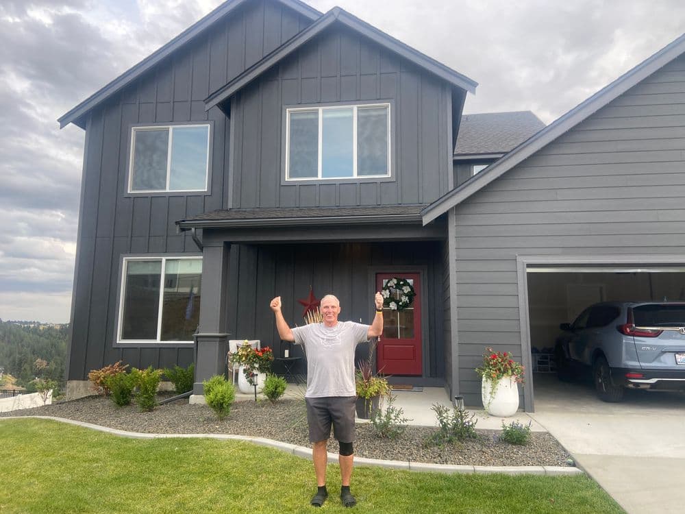 Man celebrating in front of a modern gray two-story house with a red door and landscaped yard.