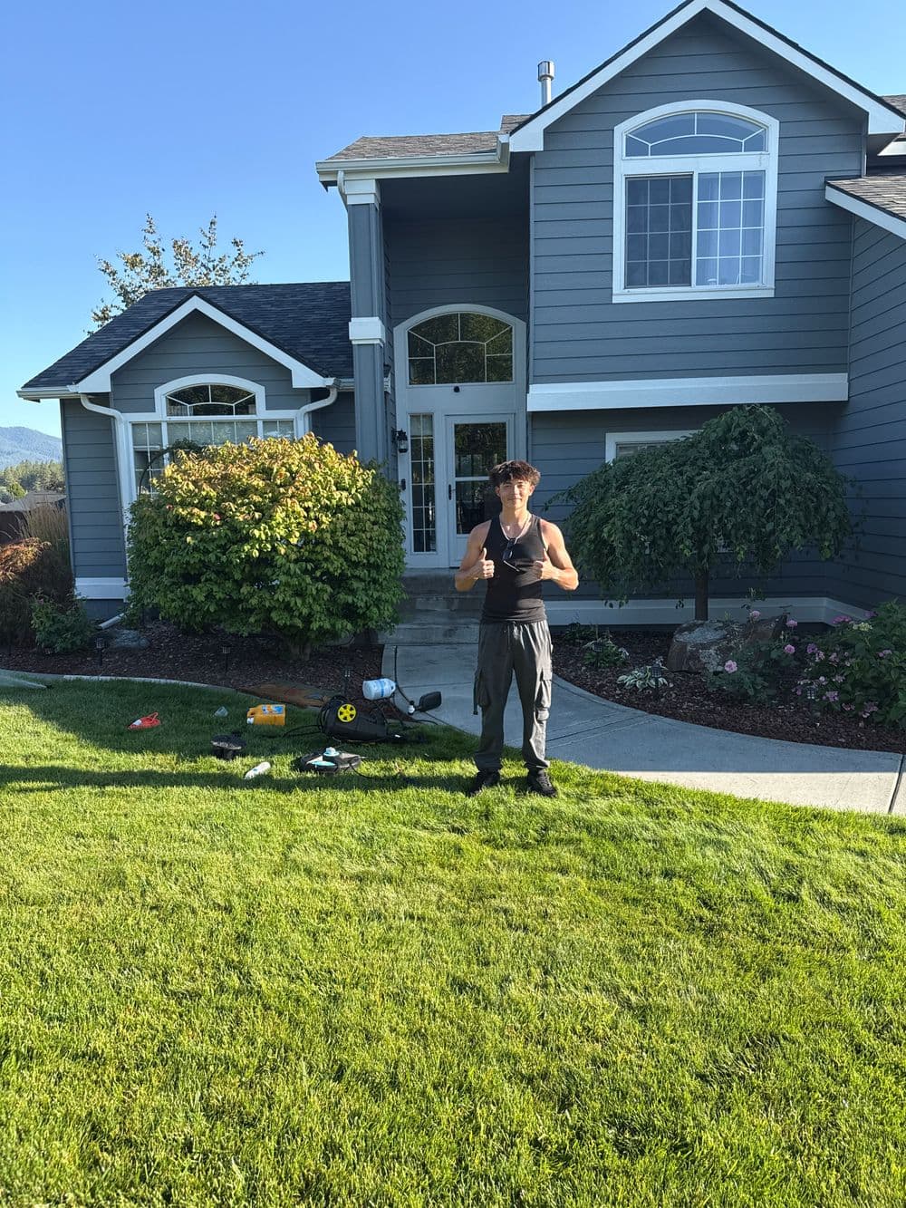 Person standing in front of a gray house on a sunny day, with a well-kept lawn and garden.