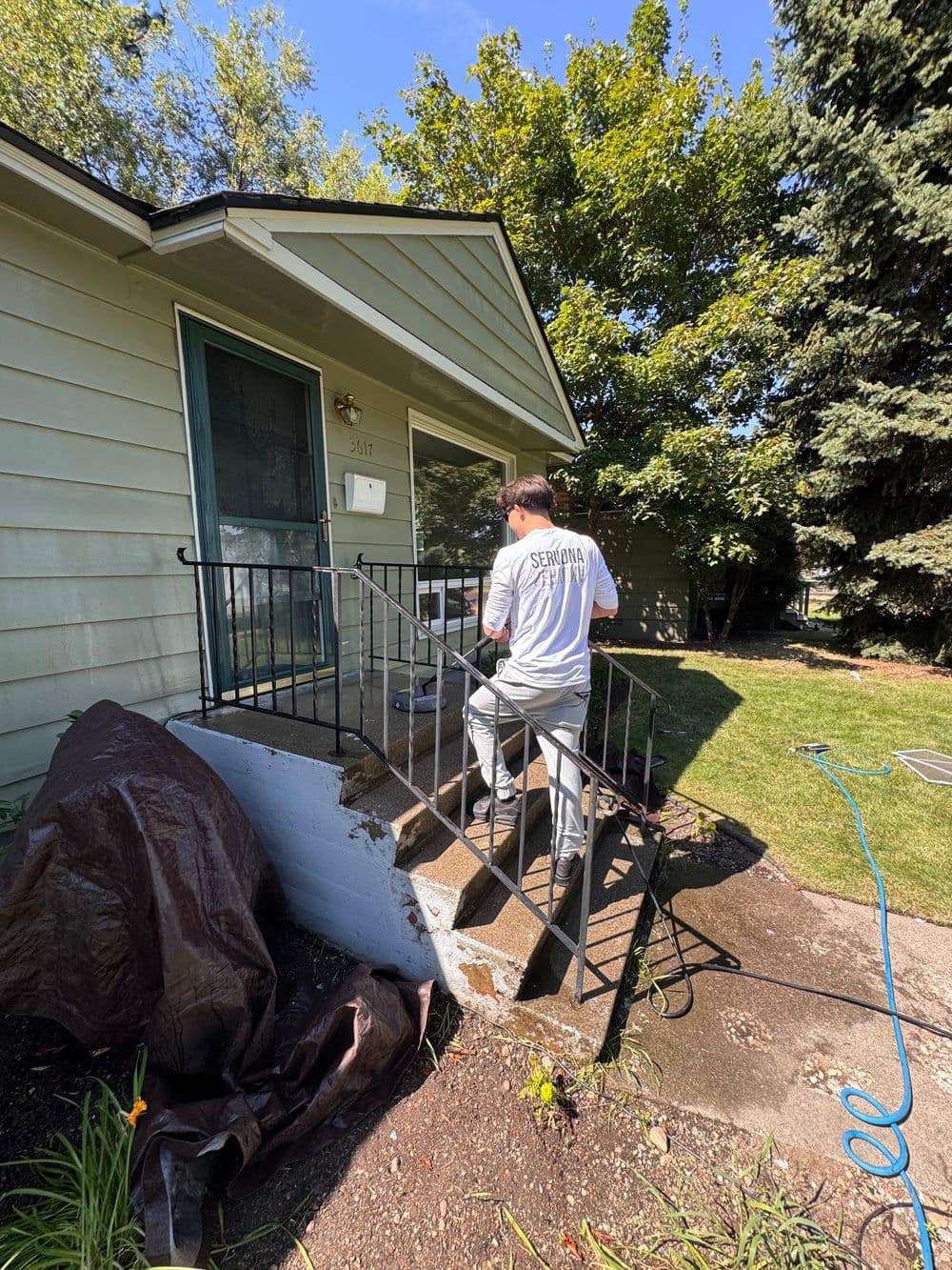 Worker cleaning outdoor stairs of a house on a sunny day, surrounded by greenery.