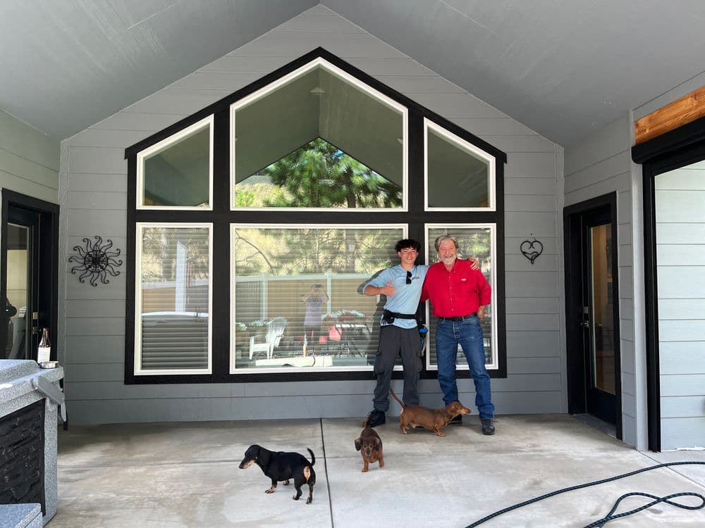 Two men pose outside a modern home with large windows, accompanied by two dogs.