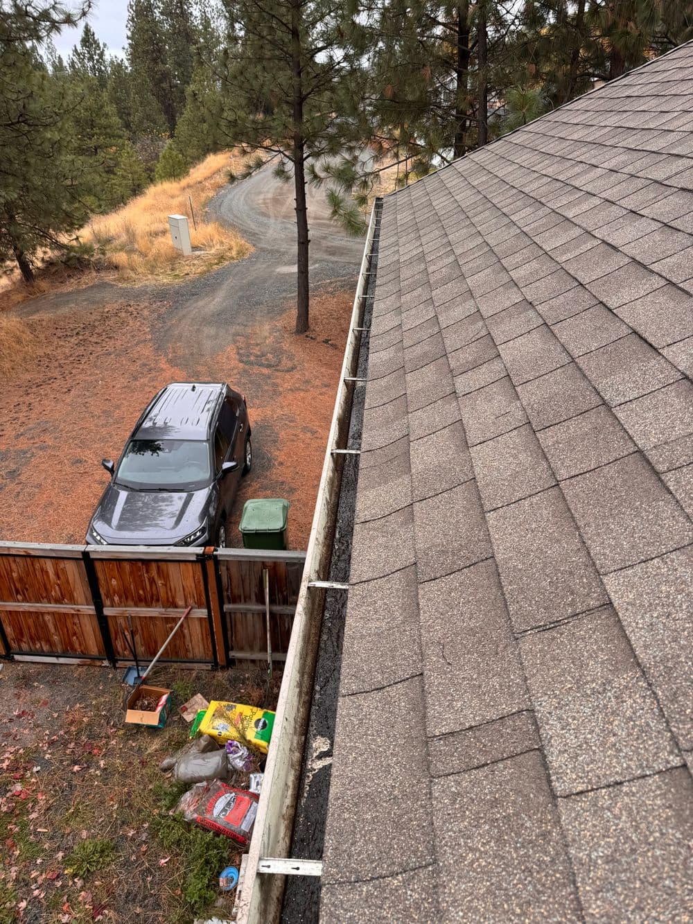 Roofline view with shingles, nearby vehicle, trash cans, and gravel road in a wooded area.