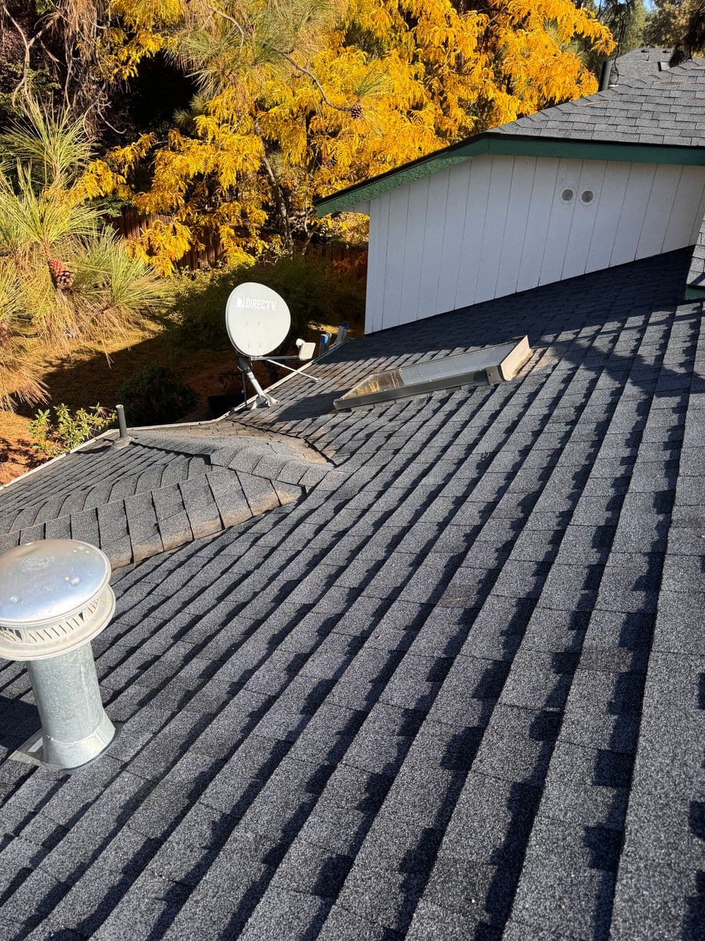 Roof with gray shingles, ventilation pipe, skylight, and satellite dish surrounded by autumn foliage.