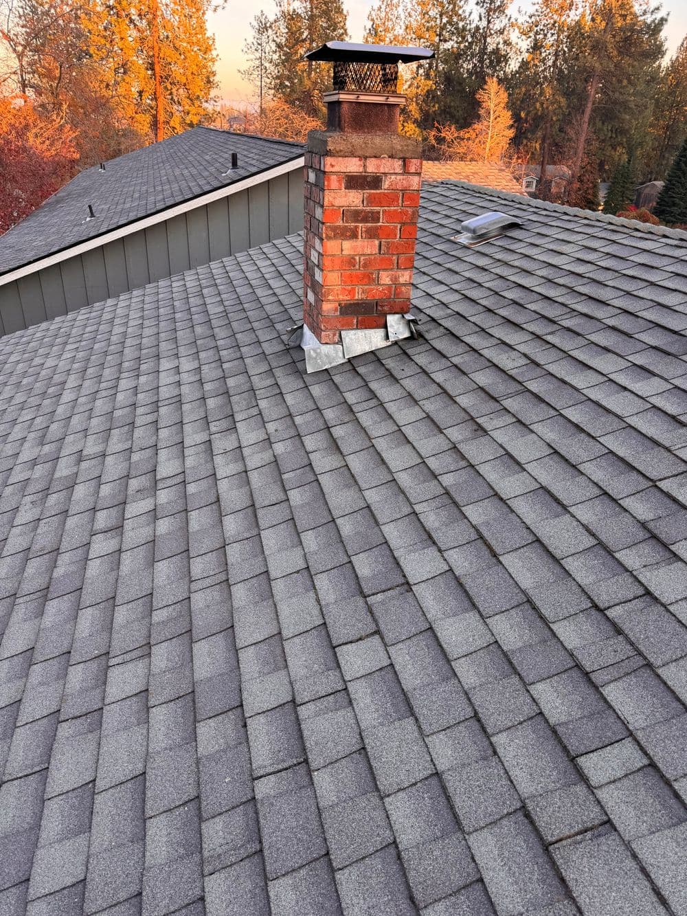 Roof with gray shingles and a brick chimney, surrounded by autumn trees.
