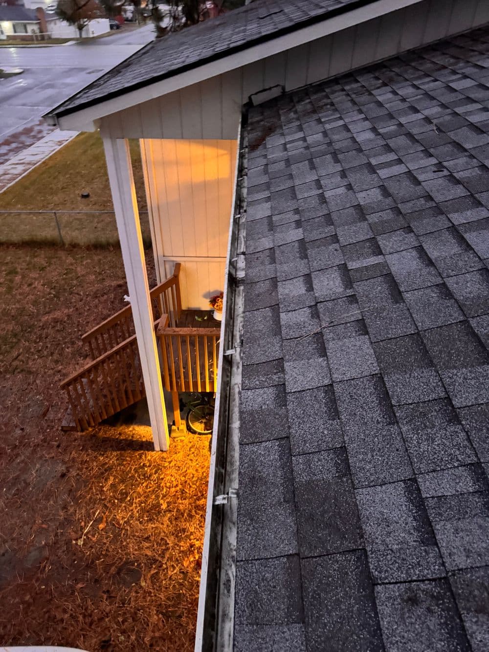 Aerial view of a house corner with a lit porch and wet asphalt outside on a cloudy day.