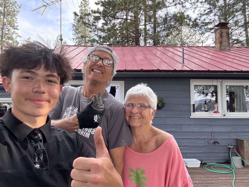 A young man gives a thumbs up with an older couple at a cozy cabin in the woods.